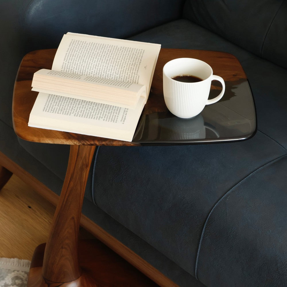 A close-up of a walnut wood side table with a glossy black resin top, featuring a coffee cup and an open book on a navy sofa.