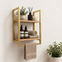 Gold wall mounted 2-tier bathroom shelf with towel bar holding bathroom bottles, towels, and decor.