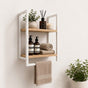 White wall mounted 2-tier bathroom shelf with towel bar holding bathroom bottles, towels, and decor.