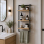 Black wall mounted 3-tier bathroom shelf with towel bar and wood-look shelves beside a modern vanity.