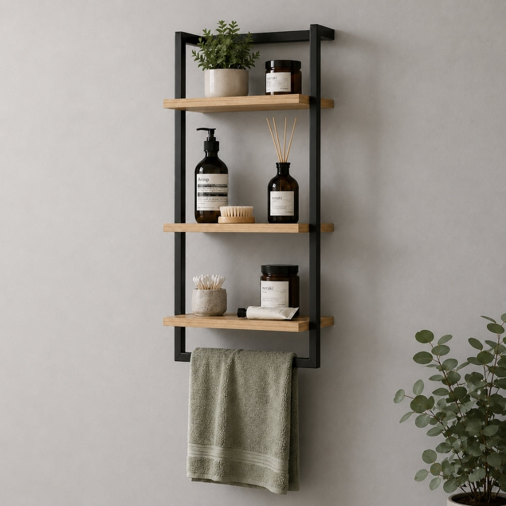 Black wall mounted 3-tier bathroom shelf with towel bar holding toiletries, plant, jars, and green towel.