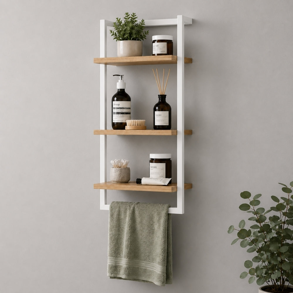 White wall mounted 3-tier bathroom shelf with towel bar holding toiletries and a green towel.