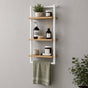 White wall mounted 3-tier bathroom shelf with towel bar holding toiletries and a green towel.