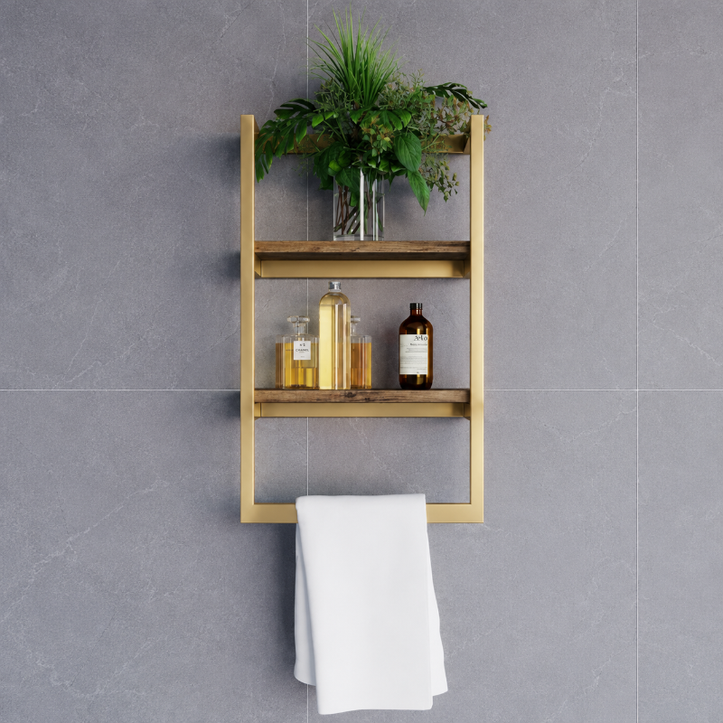 A gold-framed three-tier wooden wall shelf mounted on a gray tiled wall, styled with a green plant, amber bottles, and a white towel hanging from the bottom bar.