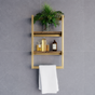 A gold-framed three-tier wooden wall shelf mounted on a gray tiled wall, styled with a green plant, amber bottles, and a white towel hanging from the bottom bar.