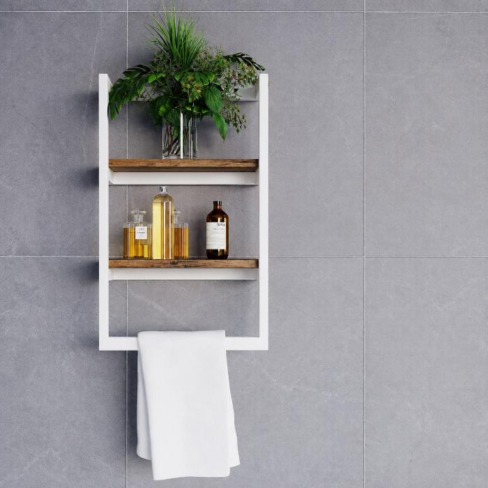 A white wall-mounted bathroom shelf with two wooden shelves and a towel bar, styled with a green plant, amber bottles, and a white towel against a gray tiled wall.
