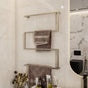 Wall-mounted towel holder in Satine Nickel finish, installed in a modern bathroom. The holder features a geometric design with multiple tiers for hanging towels. The backdrop includes a large bathtub, light-colored tiles, and a sleek toilet area, complementing the minimalist aesthetic.