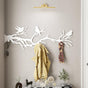 A white metal wall-mounted coat rack featuring birds perched on tree branches. The design showcases bird silhouettes sitting on elegant branches, providing both a functional hanging solution and a decorative touch. A beige coat and a patterned scarf hang from the hooks, while the scene is enhanced by minimalist decor elements below the rack.